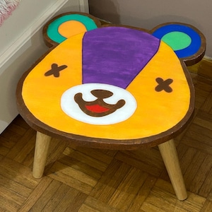 Può includere: Uno sgabello in legno con un design a forma di orso. Lo sgabello presenta un viso giallo con un muso bianco, occhi marroni a forma di croce e una bocca rossa. Le orecchie sono verdi, arancioni e blu. Lo sgabello ha tre gambe di legno.