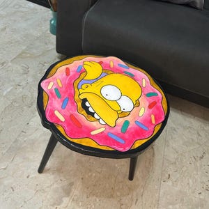 Op de afbeelding: Een ronde bijzettafel met zwarte poten, met een roze donutontwerp. De donut heeft kleurrijke hagelslag en een cartoongezicht. De tafel heeft een diameter van ongeveer 46 cm.