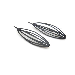 May include: Two pairs of black, leaf-shaped earrings. Each earring features a series of parallel, curved lines, creating an open, airy design. The earrings are set against a clean white background, highlighting their sleek, modern aesthetic.