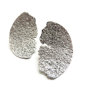 Puede incluir: Un par de pendientes de plata con una forma orgánica texturizada. Los pendientes son asimétricos y tienen un acabado martillado rugoso.