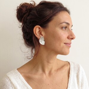 May include: A silver earring with a semi-circular shape and a textured, organic surface. The earring features a matte finish and a curved edge, positioned on the earlobe to display its distinct, leaf-like pattern and metallic sheen.