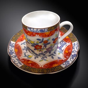 Puede incluir: Un juego de taza y platillo de cerámica blanca con un diseño floral. La taza y el platillo presentan un fondo blanco con patrones florales rojos, naranjas, azules y amarillos. El borde de la taza y el platillo tiene un borde decorativo.