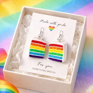 May include: A pair of earrings shaped like rainbow-layered cake slices, presented in a white gift box with a card that reads "Made with pride" and "For you, something special." The earrings have a silver hook.