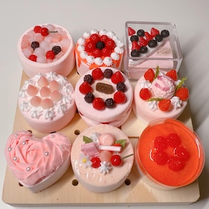 May include: A collection of nine miniature cakes and desserts arranged on a wooden pegboard. The items feature various toppings including red cherries, strawberries, blackberries, white frosting swirls, and small decorative elements in shades of pink.