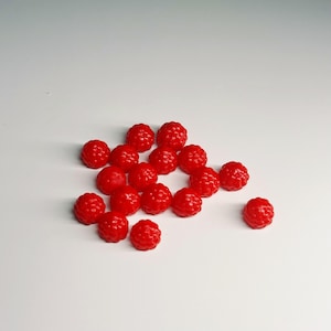 May include: A scattering of small, bright red, raspberry-shaped sweets. The sweets are arranged on a white surface, with varying sizes and textures. The image is well-lit, highlighting the vibrant colour and details of each piece.