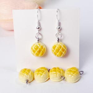 May include: Silver dangle earrings and matching stud earrings featuring a yellow, diamond-patterned design. The earrings are shaped like a small, round pastry. The earrings are displayed on a white background.