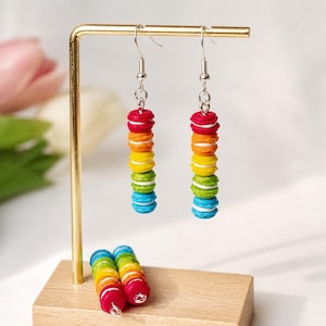 May include: A pair of colorful, stacked macaron earrings in a rainbow of colors: red, orange, yellow, green, and blue. The earrings hang from silver hooks and are displayed on a gold stand. Two additional macaron stacks rest on a wooden base.