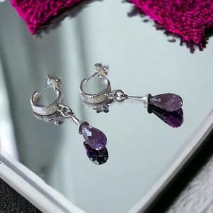 May include: A pair of silver hoop earrings with a dangling amethyst teardrop gemstone. The hoops are textured with a floral design.