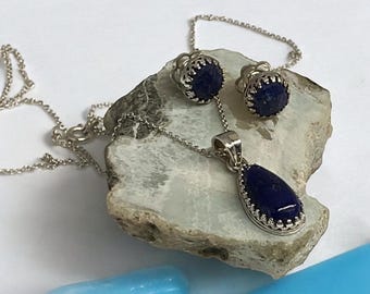 Puede incluir: Un conjunto de collar y pendientes de plata con un colgante de lapislázuli en forma de lágrima y piedras redondas de lapislázuli. Las joyas se muestran sobre una roca gris.