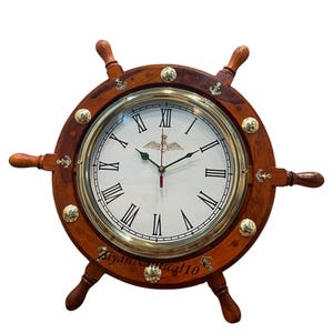 Puede incluir: Reloj de pared con temática náutica en forma de timón de barco. La esfera blanca muestra números romanos negros, con un bisel dorado y un marco de madera marrón oscuro. Se aprecian detalles decorativos de latón y anclas.
