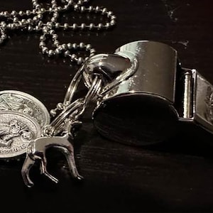 May include: A silver-colored whistle pendant necklace with a ball chain. The necklace features two oval pendants with a religious figure and a small deer charm. The items are displayed on a dark surface.