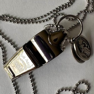 May include: A silver-colored whistle pendant with a ball chain necklace. The whistle has the text "THE ACME THUNDERER MADE IN ENGLAND". A small round charm with the text "ST. FRANCIS" is attached.