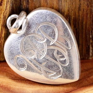 Può includere: Ciondolo a forma di cuore in argento con monogramma inciso. Il ciondolo ha un anello nella parte superiore per attaccarlo a una catena.