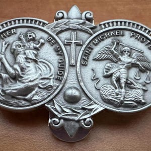 May include: Silver-toned metal religious pendant featuring three circular medallions with images of Saint Christopher, Saint Michael, and a cross. The Saint Christopher medallion has the text "St. Christopher Be My Guide". The Saint Michael medallion has the text "Saint Michael Protect Us".