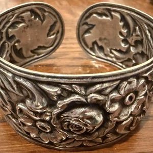 May include: Silver-toned cuff bracelet with an open design. The bracelet features detailed floral and leaf patterns, including roses and other flowers. The ends of the bracelet have a decorative oval shape.