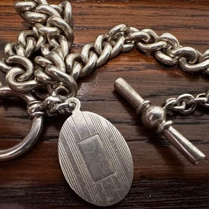 May include: Close-up of a silver pocket watch chain with a decorative oval fob and a T-bar. The chain is made of thick, interlocking links. The fob has a rectangular design. The items are on a dark wood surface.