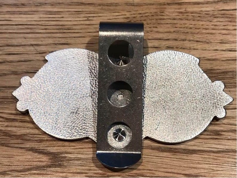 May include: Silver-toned metal belt buckle with a decorative design. The buckle has a clip on the back for attaching to a belt.