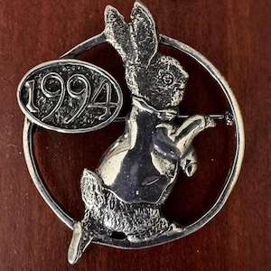May include: A silver-toned bunny brooch with the year "1994" on an oval plaque. The bunny is dressed in a vest and holding a small object. The brooch is set within a circular frame.