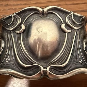 May include: Ornate silver-toned cuff bracelet with an Art Nouveau design. The bracelet features a central oval shape surrounded by flowing, stylized floral and leaf motifs. The metal has a slightly aged appearance.