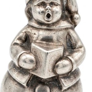 May include: A silver-toned metal charm shaped like a figure wearing a cap and coat, holding an open book. The figure has a round base with two circular buttons and a small loop at the top for attachment, featuring a detailed face with an open mouth.