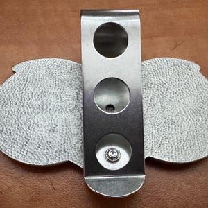 May include: A silver metal belt buckle with a textured surface and three round holes. The buckle is designed to be attached to a belt.