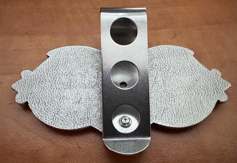 May include: A silver metal belt buckle with a textured surface and three round holes. The buckle is designed to be attached to a belt.