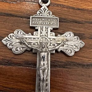 May include: Ornate silver-toned crucifix with the figure of Jesus Christ. The cross features intricate detailing and the inscription "JESUS NAZARENUS REX JUDAEORUM". The crucifix has a decorative top and a loop for hanging.