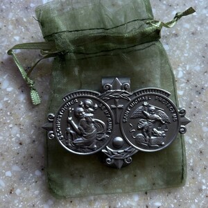 May include: A silver-toned metal money clip with two round medallions. The left medallion features Saint Christopher carrying a child, with the text "Saint Christopher Be My Guide". The right medallion features Saint Michael slaying a dragon, with the text "Saint Michael Protect Us". The money clip is in a green organza bag.