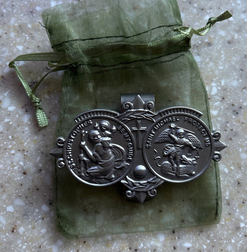May include: A silver-toned metal money clip with two round medallions. The left medallion features Saint Christopher carrying a child, with the text "Saint Christopher Be My Guide". The right medallion features Saint Michael slaying a dragon, with the text "Saint Michael Protect Us". The money clip is in a green organza bag.