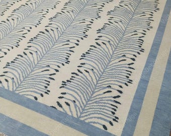 May include: A rectangular rug with a cream-colored background and a repeating pattern of light blue leaf-like shapes. The rug has a light blue border and a cream-colored inner border. The design is symmetrical and the rug appears to be made of a woven material.
