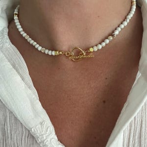 May include: A white beaded necklace with gold-coloured accents and a gold-coloured clasp. The necklace features small, round white beads with subtle brown marbling. The clasp is a decorative toggle style.
