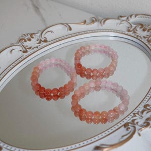 May include: Three circular bracelets made of smooth, round, translucent beads in varying shades of pink and peach. The beads exhibit a subtle gradient effect, transitioning from lighter, pale pink tones to deeper, more saturated coral-orange hues.