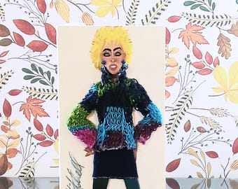 May include: A greeting card featuring a stylized illustration of a person with bright yellow hair, wearing a textured, multi-colored knit sweater in shades of blue, green, and purple, a dark blue skirt, dark leggings, and blue heels, with a signature.