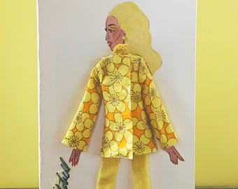 May include: A greeting card featuring a fashion illustration of a figure with blonde hair wearing a yellow and orange floral print jacket, solid yellow trousers, and yellow high-heeled shoes. The name Willie Mitchell is written in cursive script.