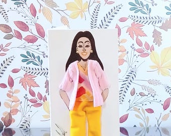 May include: A greeting card featuring a three-dimensional illustration of a person with long dark hair, wearing a light pink cardigan over a patterned orange and pink top, paired with bright yellow cuffed trousers and matching yellow shoes.