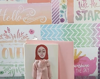 May include: A greeting card featuring a paper doll illustration of a person with red hair, wearing a light pink jacket, a tan skirt, and patterned leggings. The card is light pink and stands upright, displaying a detailed fashion-focused design.