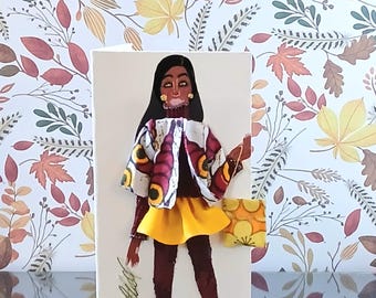May include: A greeting card featuring a collage of a figure with long black hair, wearing a dark brown turtleneck, a yellow skirt, and a patterned jacket with red, yellow, and black circular motifs. A small yellow floral bag is attached to the side.