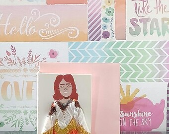 May include: A greeting card featuring a paper cutout of a person with red hair, wearing a white textured top with brown and orange accents, a yellow skirt, yellow leggings, and white shoes. The card is set against a background with floral patterns.
