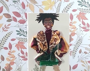 May include: A mixed-media art piece featuring a doll with dark skin and spiky black hair, wearing a high-neck maroon top, a patterned jacket with gold and brown tones, a green skirt, and mesh leggings with small studs, finished with green shoes.