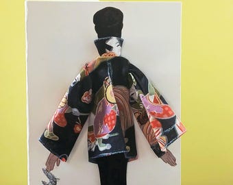 May include: A decorative card featuring a three-dimensional paper doll figure wearing a black kimono with a colorful floral and geometric pattern. The figure has dark hair styled in a bun and is holding a silver glittery accent on the left side.