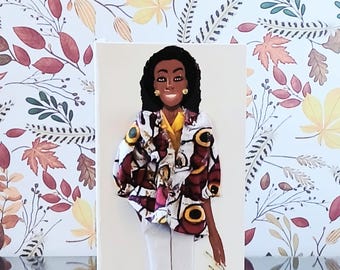 May include: A decorative wall art piece featuring a three-dimensional figure of a person with dark hair, wearing a yellow shirt under a patterned jacket with burgundy, white, and gold circular motifs, paired with white trousers and gold earrings.