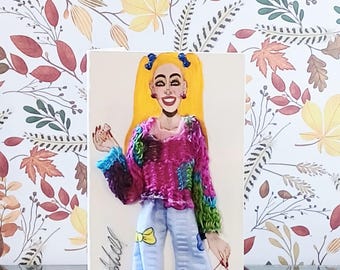 May include: A greeting card featuring a hand-drawn illustration of a person with bright yellow hair in pigtails, wearing a textured multi-colored sweater and light blue patterned pants. The person holds a pink handbag and has the name Willie Mitchell signed.