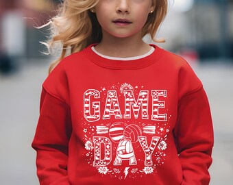 May include: A red sweatshirt with the words "GAME DAY" in white, along with volleyball graphics. The sweatshirt is worn by a young person with blonde hair, standing in front of a blurred background.