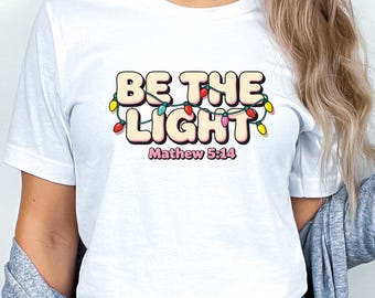 May include: White t-shirt with the text "BE THE LIGHT" in a bubble font, decorated with colorful string lights. The text "Mathew 5:14" is below the main text. The shirt is worn by a person.