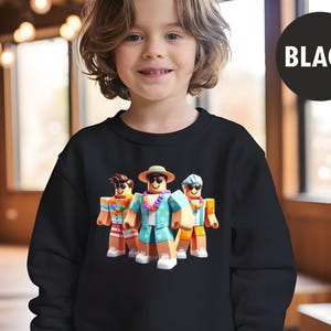 Puede incluir: Sudadera negra con un gr&aacute;fico de tres figuras en bloques con atuendos tropicales, que incluyen gafas de sol, collares de flores y una tabla de surf. La palabra "BLACK" est&aacute; en un c&iacute;rculo negro en la esquina superior derecha.
