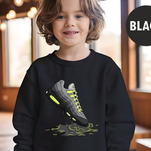 May include: Black sweatshirt featuring a graphic of a gray and black sneaker with neon green accents. The sneaker is depicted with a splash effect. A black circle with the word "BLACK" is in the upper right corner.