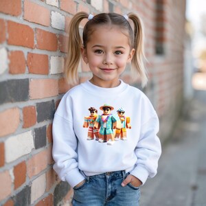 Puede incluir: Una sudadera blanca con un gr&aacute;fico colorido de tres personajes de dibujos animados con ropa de playa. Los personajes llevan gafas de sol, sombreros y camisas hawaianas. La sudadera la lleva una ni&ntilde;a peque&ntilde;a con coletas rubias y vaqueros.