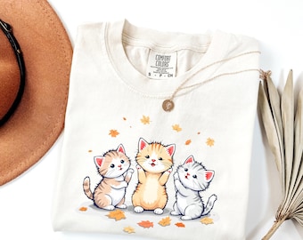May include: Cream-colored Comfort Colors sweatshirt featuring a graphic of three cartoon kittens with autumn leaves. The kittens are in shades of orange, tan, and gray. A gold necklace with a pendant is draped on the shirt.