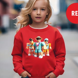 Puede incluir: Una sudadera roja con cuello blanco, que muestra un gr&aacute;fico de tres personajes de dibujos animados con atuendos de vacaciones. La palabra "RED" est&aacute; en un c&iacute;rculo rojo en la parte superior derecha.