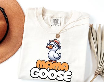 May include: Cream-colored t-shirt featuring a cartoon goose wearing glasses and a hat. The text "MAMA GOOSE" is printed in orange and white. A gold necklace is draped across the shirt. A brown hat and dried plant are in the background.